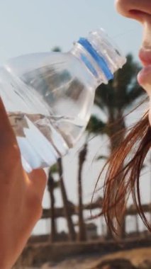 Yakın plan, güneş gözlüklü ve güneş şapkalı kadın sıcak bir yaz gününde plastik bir şişeden su içer..