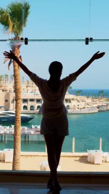 Otel panoramik penceresinin arka planına uzanmış yatlı luguna ya da marinaya bakan kadın silueti..