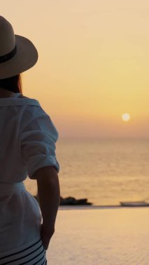 Yazlık kıyafetleri ve güneş şapkası takmış, havuzda durup güneşin doğuşunu seyreden genç bir kadın. Yaz. Deniz kıyısındaki tatil. seyahat kavramı