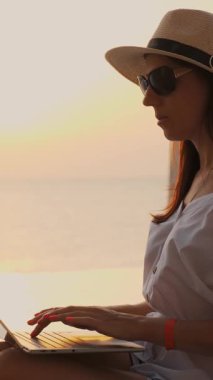 Güneş gözlüklü ve güneş şapkalı genç bir kadın, dizüstü bilgisayar kullanıyor, havuzda oturuyor, denizin üstünde güneşin doğuşuna karşı. Serbest çalışan, uzaktan kumandalı. Denizde yaz tatili