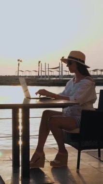 Yazlık giysiler, güneş gözlüğü ve güneş şapkası takan genç bir kadın gün batımında ya da gün doğumunda deniz kenarında tek başına gezinti masasında oturuyor. Serbest çalışan, uzaktan kumandalı. Denizde yaz tatili