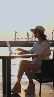 Yazlık giysiler, güneş gözlüğü ve güneş şapkası takan genç bir kadın gün batımında ya da gün doğumunda deniz kenarında tek başına gezinti masasında oturuyor. Serbest çalışan, uzaktan kumandalı. Denizde yaz tatili