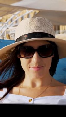 Güneş gözlüklü ve güneş şapkalı genç bir kadın, dizüstü bilgisayar kullanıyor, deniz kenarında, gün batımında ya da gün doğumunda bir yerde yatıyor. Serbest çalışan, uzaktan kumandalı. Denizde yaz tatili