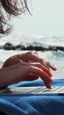 Yakın plan, parlak turuncu tırnaklı kadın elleri dizüstü bilgisayarda, deniz kenarında, sıcak yaz gününde bir şeyler yazıyor. Serbest çalışan, uzaktan kumandalı. Denizde yaz tatili