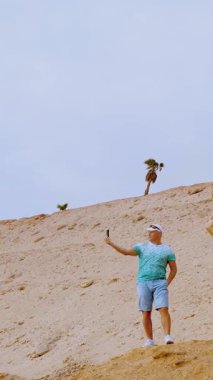 Yakışıklı atletik adam cep telefonuyla konuşuyor, selfie çekiyor, çevrimiçi video yayınına öncülük ediyor. Büyük taşların yanında, çöl zeminine karşı, tek palmiyeli. blog yazarı bir video kaydediyor ,