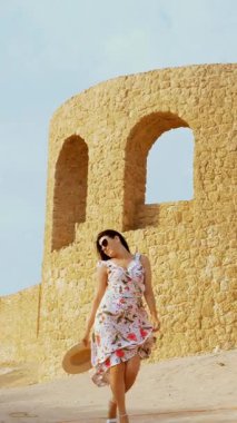 Gülümseyen, güzel genç bir kadın, güneş gözlüğü ve açık yaz elbisesi içinde, rüzgarda dalgalanan, çölün ortasında duruyor, mimari bir binanın arka planında, sıcak bir yaz gününde.