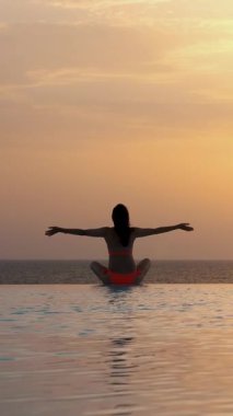 Mayolu bir kadın silueti, yoga yapıyor, meditasyon yapıyor, elleri havada dinleniyor, güneş doğarken panoramik deniz manzaralı açık sonsuzluk havuzunun kenarında..