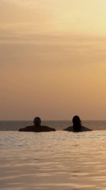 Siluetler, birbirine aşık çiftler, kadın ve erkek, suda dinleniyorlar, güneş doğarken panoramik deniz manzaralı sonsuz havuzun kenarında. balayı, seyahat ve tatil konsepti