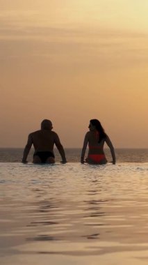 Siluetler, birbirine aşık çiftler, kadın ve erkek, suda dinleniyorlar, güneş doğarken panoramik deniz manzaralı sonsuz havuzun kenarında. balayı, seyahat ve tatil konsepti
