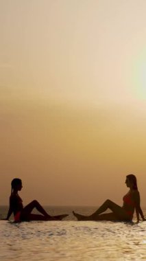 Siluetler, kadın ve genç kız, anne ve kız, güneş doğarken panoramik deniz manzaralı açık hava sonsuz havuzunun kenarına oturur ve rahatlarlar. Aile tatili kavramı