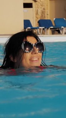 Islak, dalgalı saçlı, güneş gözlüklü, açık havada sonsuzluk havuzunda yüzen yakın plan, gülümseyen bir kadın. seyahat ve tatil kavramı