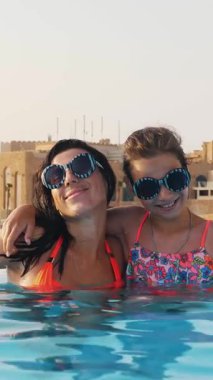 Kadın ve genç kız, anne ve kız, komik, tıpatıp aynı, yuvarlak güneş gözlükleriyle, rahatlayın, lüks otel manzaralı açık havuzda eğlenin. Aile tatili kavramı
