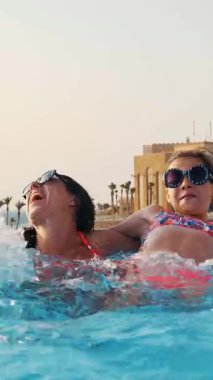 Kadın ve genç kız, anne ve kız, komik, tıpatıp aynı, yuvarlak güneş gözlükleriyle, rahatlayın, lüks otel manzaralı açık havuzda eğlenin. Aile tatili kavramı