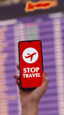 Yaklaş, kadın, koruyucu maskeli, uçak ikonlu akıllı telefon, dur işareti, ekranda, kalkış terminalinde, havaalanında uçuş bilgi panosunun önünde.