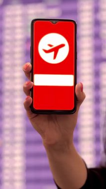 Yaklaş, kadın, koruyucu maskeli, akıllı telefonu uçak simgesiyle tutuyor, işaret veriyor, ekranda, kalkış terminalinde, havaalanında, uçuş bilgi panosunun önünde.