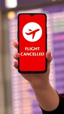 Yaklaş, kadın, koruyucu maskeli, uçak ikonlu akıllı telefonu elinde, uçuş iptal işareti, ekranda, havaalanındaki uçuş bilgi panosunun önünde.
