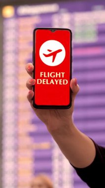 Yaklaş, kadın, koruyucu maskeli, uçak ikonlu akıllı telefonu tutuyor, uçuş gecikmeli işareti, ekranda, kalkış terminalinde, havaalanında uçuş bilgi panosunun önünde.