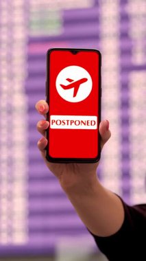 Yaklaş, kadın, koruyucu maskeli, akıllı telefonu uçak simgesiyle tutuyor, ertelenmiş işareti, ekranda, kalkış terminalinde, havaalanında, uçuş bilgi panosunun önünde.