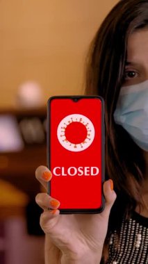 Kadın, koruyucu maskeli, elinde COVID-19 simgesi olan akıllı bir telefon tutuyor, mesaj - kapalı, ekranda, havaalanındaki boş kayıt gişelerinin önünde.