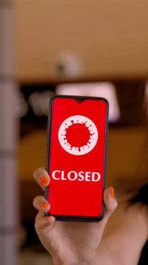 Kadın, koruyucu maskeli, elinde COVID-19 simgesi olan akıllı bir telefon tutuyor, mesaj - kapalı, ekranda, havaalanındaki boş kayıt gişelerinin önünde.