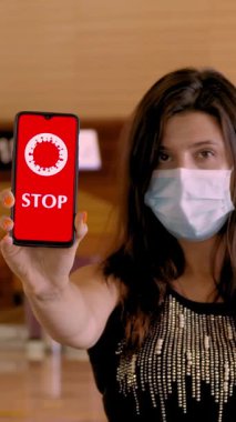 Kadın, koruyucu maskeli, elinde COVID-19 simgesi olan bir akıllı telefon tutuyor, mesaj yazıyor - ekranda, havaalanındaki boş kayıt gişelerinin önünde,