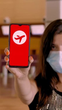 Koruyucu maskeli bir kadın, elinde uçak ikonu, mesaj, uçuş iptal edildi, ekranda, havaalanındaki boş kayıt gişelerinin önünde akıllı telefon tutuyor..