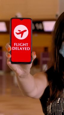 Koruyucu maskeli bir kadın, elinde uçak ikonu, mesajı olan akıllı bir telefon tutuyor. Uçuş gecikmeli, ekranda, havaalanındaki boş gişelerin önünde..