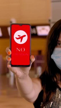Koruyucu maskeli bir kadın, elinde uçak ikonu, mesajı olan akıllı bir telefon tutuyor. Tatil yok, ekranda, havaalanındaki boş gişelerin önünde..