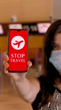 Koruyucu maskeli bir kadın, elinde uçak ikonu, mesajı olan akıllı telefonu havaalanındaki boş gişelerin önünde, ekranda gezmeyi durduruyor..