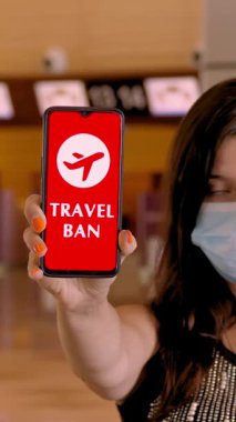 Koruyucu maskeli bir kadın, elinde uçak ikonu, mesaj, seyahat yasağı olan akıllı telefonu havaalanındaki boş gişelerin önünde, ekranda tutuyor..