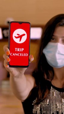 Kadın, koruyucu maskeli, elinde uçak ikonu, mesaj içeren akıllı telefonu var. Havaalanındaki boş gişelerin önünde, ekranda tatil iptal edildi..