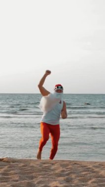 Komik Noel Baba. Noel Baba deniz kenarında, yaz tatilinde. Noel Baba dans ediyor, deniz sahilinde atlıyor, gün doğumunda. Noel Baba tişörtlü ve deniz kıyısında kırmızı taytlı..