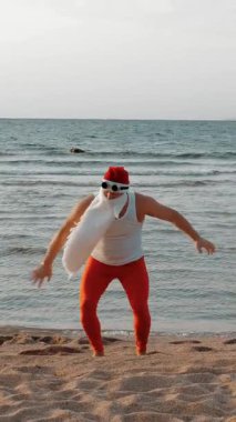 Komik Noel Baba. Noel Baba deniz kenarında, yaz tatilinde. Noel Baba dans ediyor, deniz sahilinde atlıyor, gün doğumunda. Noel Baba tişörtlü ve deniz kıyısında kırmızı taytlı..