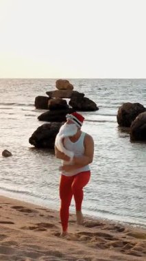 Komik Noel Baba. Noel Baba deniz kenarında, yaz tatilinde. Noel Baba dans ediyor, deniz sahilinde atlıyor, gün doğumunda. Noel Baba tişörtlü ve deniz kıyısında kırmızı taytlı..