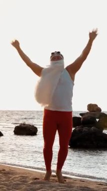 Komik Noel Baba. Noel Baba deniz kenarında, yaz tatilinde. Noel Baba dans ediyor, deniz sahilinde atlıyor, gün doğumunda. Noel Baba tişörtlü ve deniz kıyısında kırmızı taytlı..