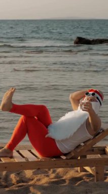 Komik Noel Baba güneşleniyor. Noel Baba sahildeki tahta şezlongda uzanıyor ve lezzetli bir kokteyl içiyor. Noel Baba yaz tatilinde, deniz kıyısında..