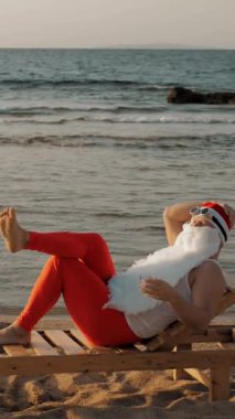 Komik Noel Baba güneşleniyor. Noel Baba sahildeki tahta şezlongda uzanıyor ve lezzetli bir kokteyl içiyor. Noel Baba yaz tatilinde, deniz kıyısında..