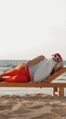 Komik Noel Baba güneşleniyor. Noel Baba, güneş gözlüklü, deniz kıyısındaki tahta şezlongda yatıyor. Uyuyor ve dinleniyor. Noel Baba yaz tatilinde, deniz kıyısında.