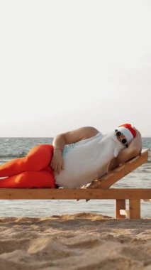 Komik Noel Baba güneşleniyor. Noel Baba, güneş gözlüklü, deniz kıyısındaki tahta şezlongda yatıyor. Uyuyor ve dinleniyor. Noel Baba yaz tatilinde, deniz kıyısında.
