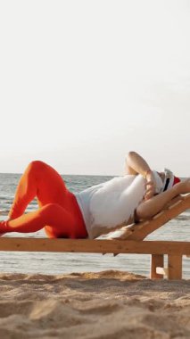 Komik Noel Baba güneşleniyor. Noel Baba, güneş gözlüklü, deniz kıyısındaki tahta şezlongda yatıyor. Uyuyor ve dinleniyor. Noel Baba yaz tatilinde, deniz kıyısında.