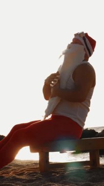 Komik Noel Baba güneşleniyor. Noel Baba sahildeki tahta şezlongda oturuyor, gün doğumunda, dinleniyor, beyaz sakalını okşuyor. Noel Baba yaz tatilinde, deniz kıyısında..