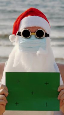 Koruyucu maskeli komik Noel Baba. Noel Baba, güneş gözlüklü, elinde yeşil bir işaret var, denizin arka planında. Coronavirus salgını sırasında Noel Baba tatili ya da deniz kıyısında tecrit..