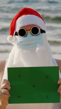 Koruyucu maskeli komik Noel Baba. Noel Baba, güneş gözlüklü, elinde yeşil bir işaret var, denizin arka planında. Coronavirus salgını sırasında Noel Baba tatili ya da deniz kıyısında tecrit..