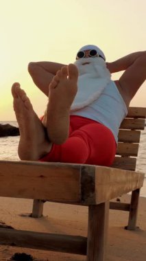 Komik Noel Baba güneşleniyor. Noel Baba, güneş gözlükleriyle, sahildeki tahta şezlongda uzanır, dinlenir. Noel Baba yaz tatilinde, deniz kıyısında..