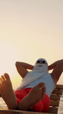 Komik Noel Baba güneşleniyor. Noel Baba, güneş gözlükleriyle, sahildeki tahta şezlongda uzanır, dinlenir. Noel Baba yaz tatilinde, deniz kıyısında..