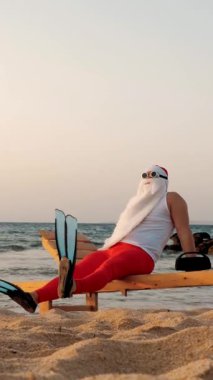 Komik Noel Baba güneşleniyor. Noel Baba, güneş gözlüğü ve palet takmış, deniz kıyısında, dinleniyor ve müzik dinliyor, dinleniyor. Noel Baba yaz tatilinde, deniz kıyısında..