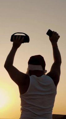 Noel Baba dans ediyor. Komik Noel Baba elinde kablosuz bluetooth hoparlörüyle gün batımına ya da gün doğumuna karşı dans ediyor. Arkadan bak. Kumsalda deniz kenarında rahatla.