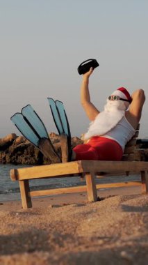 Noel Baba güneşleniyor. Komik Noel Baba, güneş gözlüğü ve palet takmış, deniz kıyısında uzanmış, elinde küçük bir müzikle müzik dinliyor, dinleniyor. Noel Baba yaz tatilinde, deniz kıyısında..