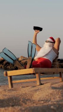 Noel Baba güneşleniyor. Komik Noel Baba, güneş gözlüğü ve palet takmış, deniz kıyısında uzanmış, elinde küçük bir müzikle müzik dinliyor, dinleniyor. Noel Baba yaz tatilinde, deniz kıyısında..