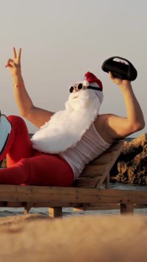 Noel Baba güneşleniyor. Komik Noel Baba, güneş gözlüğü ve palet takmış, deniz kıyısında uzanmış, elinde küçük bir müzikle müzik dinliyor, dinleniyor. Noel Baba yaz tatilinde, deniz kıyısında..